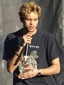 A young man with dark roots to his short bleached hair uses a cordless handheld mic to perform while wearing a black t-shirt with a colorful decal.
