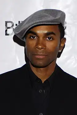 Fab Morvan wearing a dark jacket and a grey flat cap, looking directly at camera