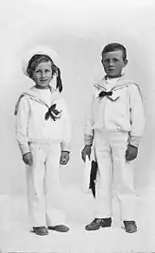 A black and white portrait of two young boys. They are standing alongside one another and are dressed in white sailor uniforms. The shorter boy on the left is smiling and wearing a cap, while the boy on the right has a blank expression and is holding a cap in his left hand.