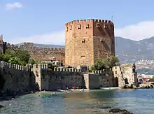 A eight-sided medieval tower built of red and yellow brick rises above a green sea in which swimmers play. Stone walls run along the shore and further up from the tower.