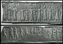 A black and white image showing two metallic plaques bearing the images of multiple persons standing in line and holding a variety of objects.