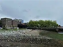 A stony beach overlooking several piers, brick buildings, and some trees