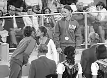 One girl in a track suit with a medal around her neck looks on as two girls congratulate each other