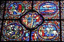 Part of a large window. It is divided into four circular panels with a quatrefoil (or four-lobed section) between them and an arched segment beneath. To the lower right, a knight, Sagittarius, is being welcomed home. At the centre a man empties a water pot, symbolising Aquarius and a two-faced figure symbolises New Year. To the upper right, are the two fish of Pisces. The other sections show each month's activities: grapevine pruning, bell-ringing and sitting by the fire.