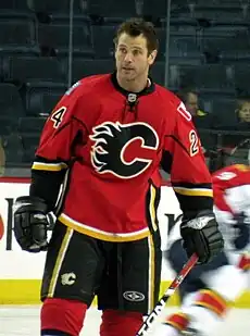 A man with short, brown hair looks to his right. He is wearing a red uniform with black pants and a black stylized C logo on his chest.