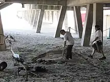 Two men, dressed in white shirts and loose pants, are seen beneath a wooden pavilion. The pavilion's legs stick out diagonally from the pavilion. One man carries a pipe which he points toward a pit in the sandy ground. The second man walks toward him.