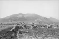 Soldiers wearing flak jackets and helmets are prone on an open field at the base of a large vegetated hill