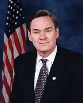 A gray-haired man in a suit sits in front of an American flag