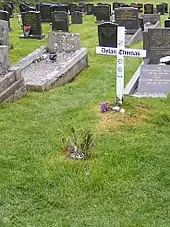 A simple white cross engraved with a memorial message to Thomas stands in a grave yard
