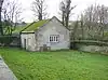 A small, simple chapel seen almost end-on in a grassy burial ground; it is built in stone with a moss-covered roof. On the end is a window with open shutters; on the front face is a door and two shuttered windows.