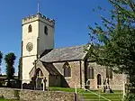 Stone building with lighter coloured square tower. In the foreground are gravestones.