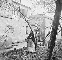 A black and white photo of a Black woman standing in front of a dilapidated and overgrown building.