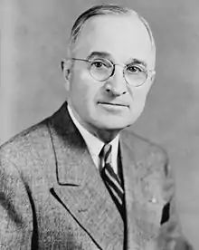 Half length portrait of a man wearing suit and tie with his head turned to looking directly forward; He has a slight smile on his face, with a receding hairline and large round glasses