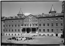 Black and white photograph of façade of building with fountain and courtyard in background. Tops of towers located behind the building are visible over its roof
