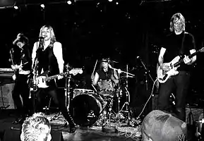 A five-piece rock band performing live on stage. A red sign behinds them reads "SPIN 20" in white lettering.