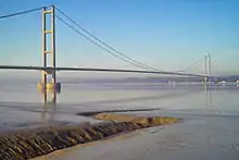 The near pier of a suspension bridge spanning calm blue waters of a wide river estuary.
