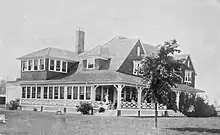 Image of the exterior of the Inverness Club House, with people seated on porch stairs, in 1910.