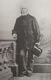 A black and white standing portrait of a white male holding a top hat