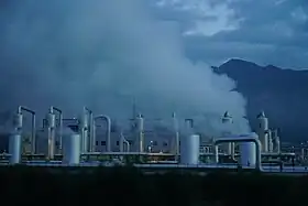 Lots of steam rises above metal pipes and vertical cylinders, with a low building and mountains in the background.