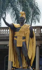 An impressive statue of a dark-skinned man, richly dressed in native garb. One arm is extended, as if in greeting, and he holds a spear, butt end grounded, in the other.