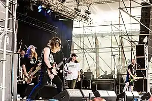 On a music stage, four men with long hair, three with electric guitars