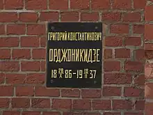 A black plaque with gold letters on a brick wall with a Russian inscription that translates as Grigory Konstantinovich Ordzhonikidze, 1886-10-28–1937-02-18