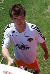 A young man with dark hair stands on a grassy surface. He is wearing a white T-shirt with black trim and orange shorts, and is smiling slightly.