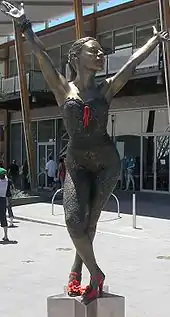 A bronze statue of Kylie, on a star-shaped pedestal, portrays her in a dancing pose. Her legs are crossed and she bends at the waist, with both arms stretched above her head. The statue stands in a public square in front of a modern glass building, and several people are walking.