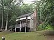 A two-story log cabin with a front porch and a forested hillside in the background.