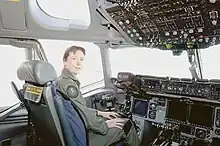 Colour photo of a woman wearing a green flight suit sitting inside an aircraft cockpit
