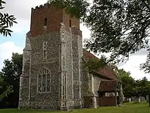 A church seen from the southwest; prominent is the west tower, its lower two stages in flint and the battlemented top stage in red brick; beyond it stretches the body of the church with its south porch