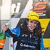 A male in his mid-20s in racing overalls sporting long hair with a yellow sponsored baseball cap on his head celebrating with a bottle of champagne in his right hand