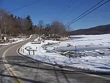 A road winding by the shore of a frozen lake and some docks in wintertime. There are telephone lines over the road, light snow on the ground, and a wooded hill on the far side of the lake at the right of the image