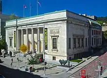 A three-story light stone building with a flat green-trimmed roof from which the Canadian, Quebec and Montreal flags are flying from poles at the center, seen from a three-quarters angle about midway up its height from across an intersection. On its front face is a recessed entry area with four fluted columns topped by Corinthian capitals. Banners to either side have the word "Chihuly", and the entablature has "Montreal Museum of Fine Arts" and "Musée de Beaux Art du Montréal in capital metal letters