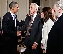 A younger African-American man shakes hands with an older man; both of them are wearing dark suits. Next to the older man is a woman wearing a white suit, facing away from the camera