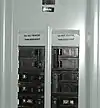 Illustration of breaker numbering in a North American type panelboard. Some labels are missing, and some are additional. The numbers on the toggles indicate the rated amperage before tripping. The top right breaker (Rated at 100&nbsp;A) feeds a sub panel.