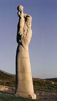 A large stone statue of a tall and slender woman by a field. The woman has long hair and wears a hat and carries a child on her shoulders.