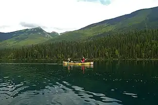Two paddlers sit in a canoe, gently paddling across the calm water. Behind them, large green mountains flank the water.
