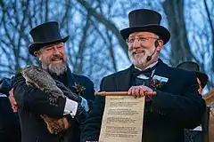 alt=Two smiling men dressed in formal attire stand beside each other. Other people and bare trees are visible in the background. The man on the left holds a groundhog in his arms. The man on the right, wearing an earpiece, holds forward an open scroll, which reads in small text:
"HEAR YE, HEAR YE, HEAR YE.
TODAY, 2-2-22, WELCOME TO PUNXSUTAWNEY TO CELEBRATE GROUNDHOG DAY, THE ONE HUNDRED THIRTY SIXTH ANNUAL TREK OF THE PUNXSUTAWNEY GROUNDHOG CLUB.
PUNXSUTAWNEY PHIL, THE SEER OF SEERS, THE PROGNOSTICATOR OF ALL PROGNOSTICATORS, WAS GENTLY LIFTED FROM HIS BURROW AT 7:25 AM, AND HELD HIGH TO SEE. HIS FAITHFUL FOLLOWERS HAD RETURNED WITH GLEE!
PLACING PHIL ON TOP OF THE STUMP, WHERE IN GROUNDHOGESE, HE DIRECTED THE PRESIDENT, JEFF LUNDY, AND THE INNER CIRCLE TO HIS PREDICTION SCROLL THAT READS:
WINTER HAS BEEN BLEAK AND DARK AND BEREFT OF HOPE. YET WINTER IS JUST ANOTHER STEP IN THE CYCLE OF LIFE. AS I LOOK OUT OVER THE FACES OF THE TRUE BELIEVERS FROM AROUND THE WORLD, I BASK IN THE WARMTH OF YOUR HEARTS. I COULDN'T IMAGINE A BETTER FATE, WITH MY SHADOW I HAVE CAST, THAN A LONG AND LUSTROUS SIX MORE WEEKS OF WINTER."
