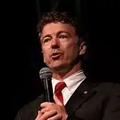 A man with brown, wavy hair wearing a suit and holding a microphone