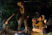 A man with a video camera kneels to film as an actor pulls a machete from the head of another actor who is sitting on the ground. Behind the cameraman, another man records sound from the scene they are filming, while a third man stands in the background.