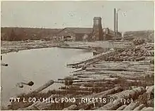 Sepia-tone photo of a pond surrounded by large logs. At the far end of the pond is a large building with a square tower and two smokestacks. Label is "T&T L CO MILL POND RICKETTS PA 1903" (i.e. Trexler and Turrell Lumber Company Mill Pond ...)
