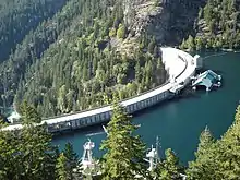 A curved concrete dam, viewed from slightly above. On the left is a steep forested valley, and on the right is blue-green water. The reservoir is almost full.