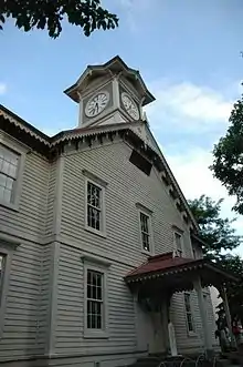 Photograph of a white wooden building against a blue sky. The building is of Western architectural design built with subtle Japanese details. A clock tower rising from the front of the building is the dominant feature.