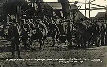 A uniformed stretcher party carry a body away from the pithead; others wait around the pithead. Wreckage from the explosion can be seen in the background.