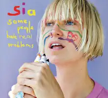 Sia stands against a light blue background, holding some markers in front of her face. There are drawings on her face. To the left of her, the name "Sia" and the title "Some People Have Real Problems" is stylized in a colorful, handwritten font.