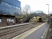 A station with two platforms and tracks. In the foreground is a 19th-century pedestrian bridge.