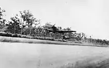 Two single engined propeller-driven monoplanes fly just above a cleared area. A thick forest is visible behind the aircraft.