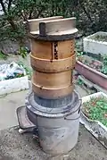 A simple hearth with a metal pan holding two wooden steaming vessels and a wooden lid used in Japan