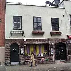 The facade of 51 Christopher Street as seen from across the street. The facade is two stories high. At the ground or first story, the facade is made of brick and consists of a wide window flanked by two archways. At the second story, there is a stucco facade with three windows, each with a flower-pot holder below.